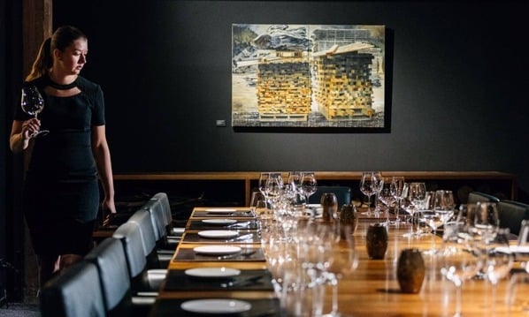 A woman stands before a table adorned with wine glasses, creating an ambience of an exclusive dining experience inside The Landscape Restaurant at Henry Jones.