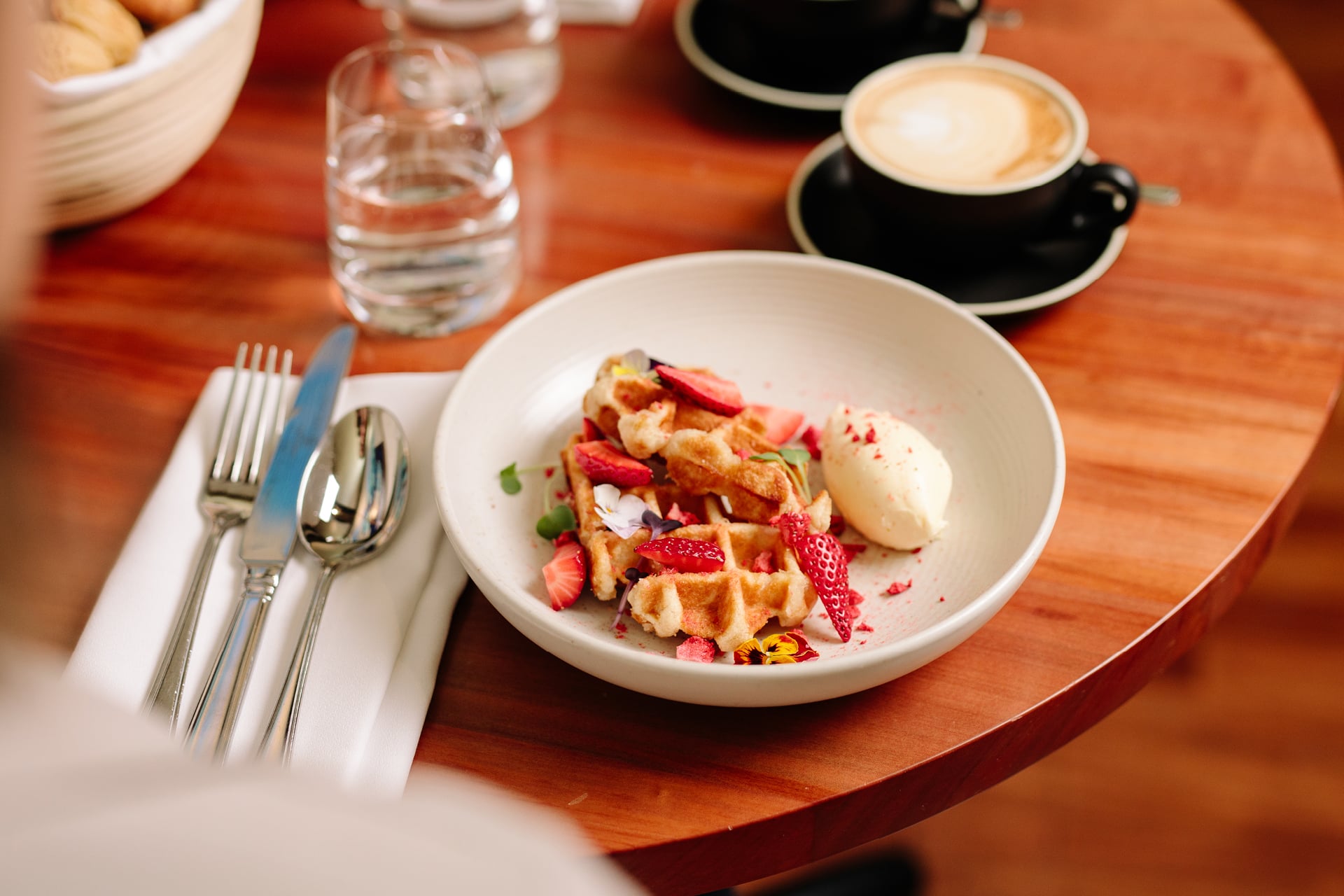 A plate of waffles topped with strawberries and ice cream, alongside a cup of water, cutleries and a cup of coffee.